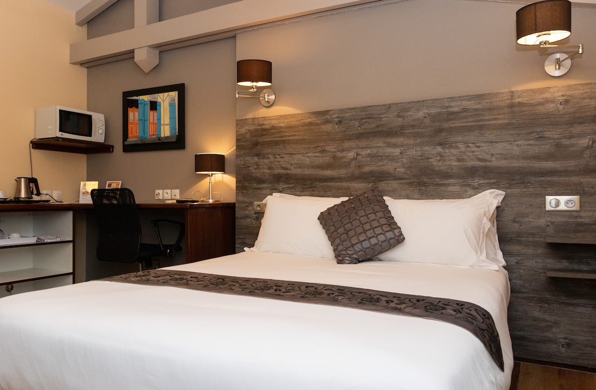 A well-appointed bedroom is showcased with a spacious double bed featuring a decorative throw pillow. A stylish headboard is complemented by modern wall sconces. A small microwave is positioned on a shelf next to a functional workspace with a chair, while artwork adorns the wall.