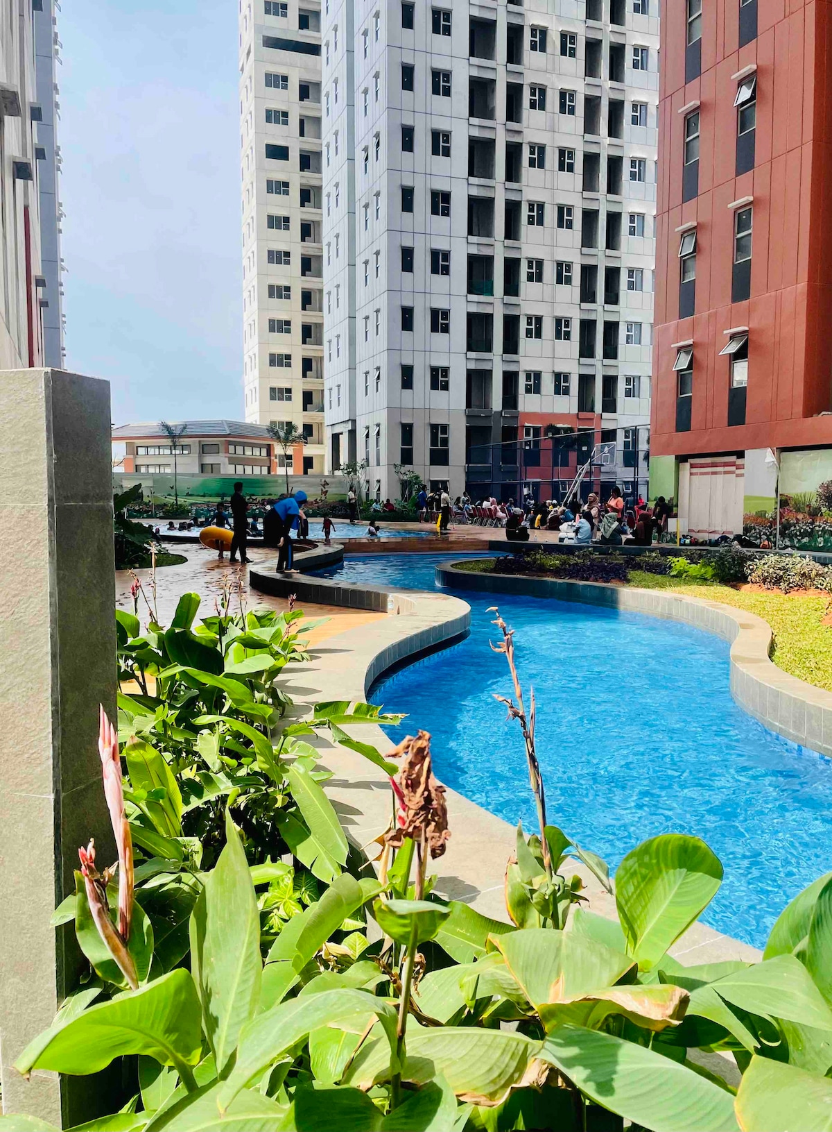 A serene outdoor pool area is surrounded by landscaped greenery and tall residential buildings. The winding pool reflects blue water, while guests can be seen enjoying the space. Tranquil pathways and flowering plants enhance the inviting environment.