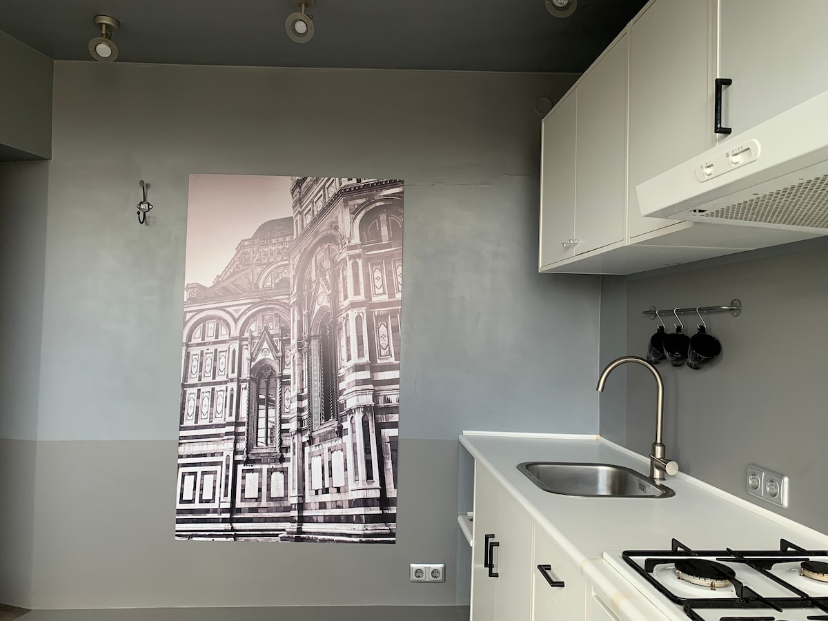 A functional kitchen space features minimalistic white cabinetry and a simple countertop. A large black and white photograph of a historic building adds visual interest to the grey wall. Cooking essentials include a stove and a sink, enhancing the practical appeal of the area.