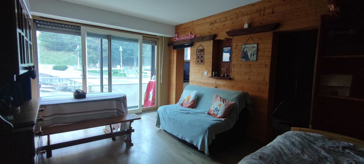 A spacious living area is featured with a light wood floor and wooden accents on the walls. A couch with decorative pillows is positioned in front of large sliding glass doors, offering natural light. A dining table is visible, complemented by a shelf displaying decorative items.