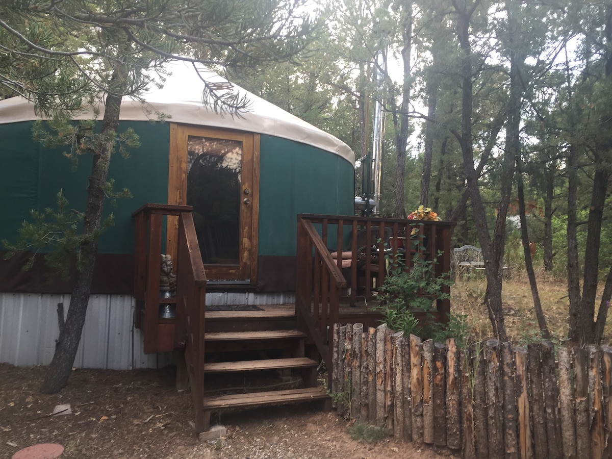 The exterior of the yurt is framed by trees, showcasing a wooden deck with steps leading to the entrance. The door is composed of natural wood and includes a small decorative arrangement. A rustic wooden fence surrounds the area, contributing to a serene retreat atmosphere.