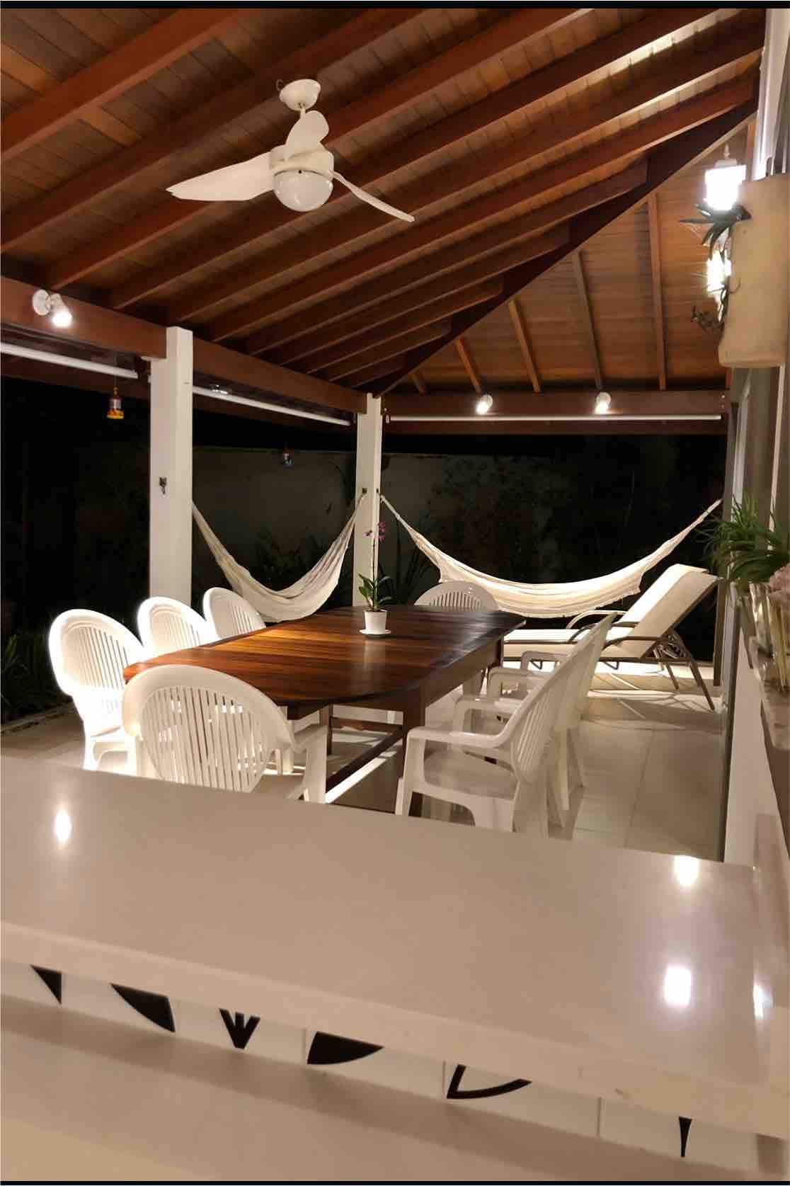 An outdoor dining area is highlighted with a wooden table surrounded by six white chairs. Two hammocks are suspended nearby under a vaulted wooden ceiling, with ambient lighting creating a relaxed atmosphere. Decorative plants are placed along the walls, enhancing the inviting space.