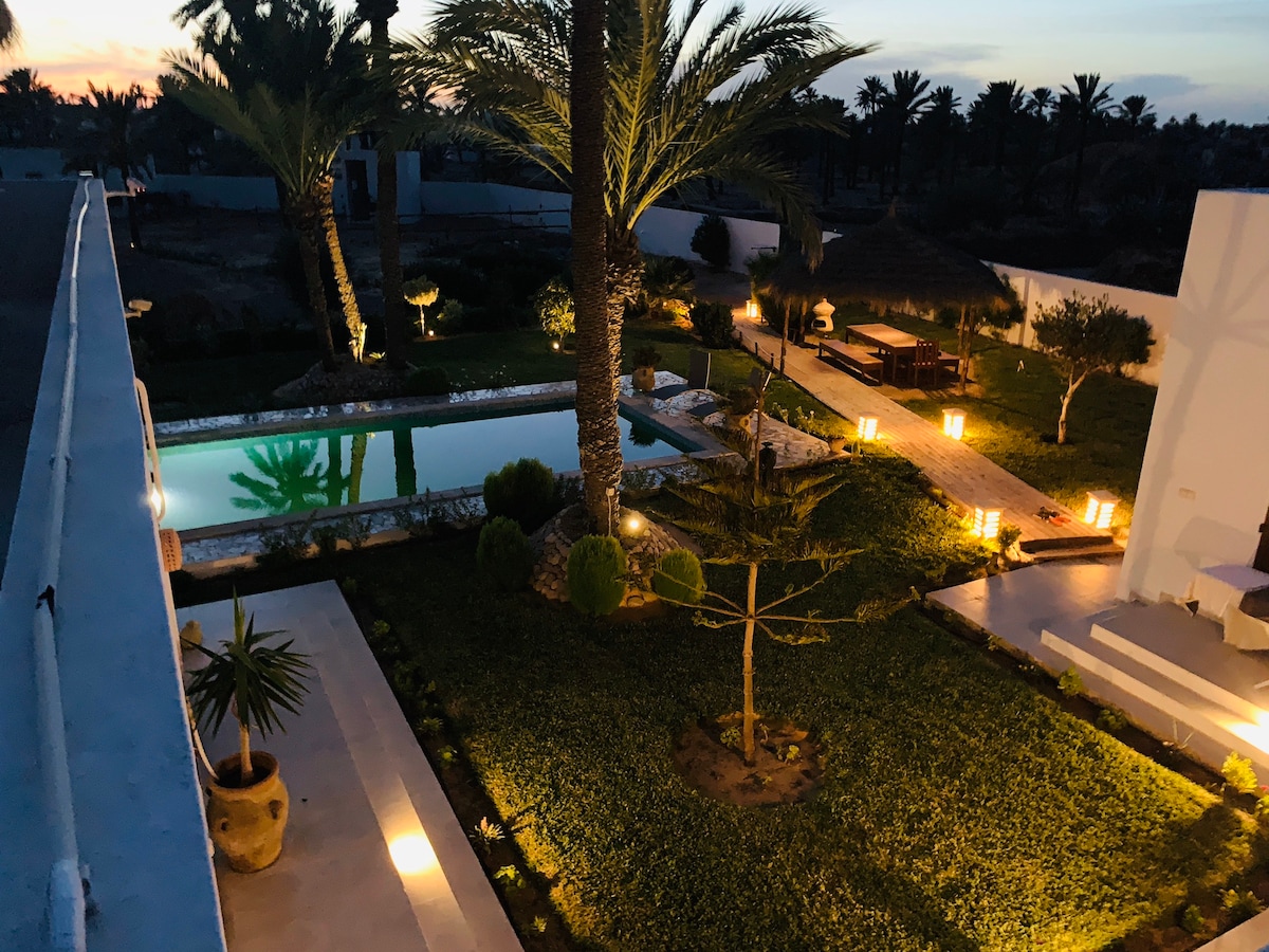 A tranquil garden setting is showcased, featuring a well-maintained pool surrounded by lush greenery and strategically placed lighting. Palm trees provide height and depth to the landscape, while various seating areas invite relaxation. The atmosphere appears calm and serene during twilight.
