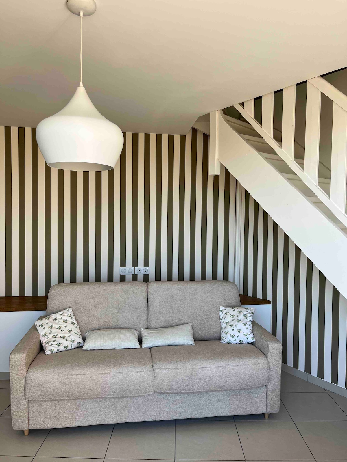 A spacious area features a neutral-toned sofa with decorative cushions, positioned against a striped wall. A pendant light extends from the ceiling, adding a contemporary touch. A staircase can be seen in the background, enhancing the room's depth.
