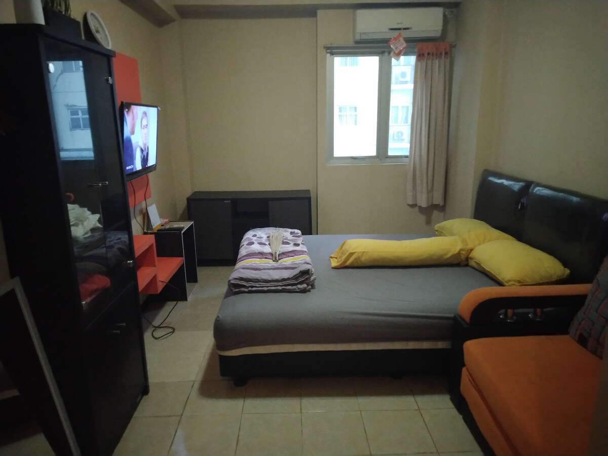 A bedroom features a spacious bed with bright yellow pillows and a light gray blanket. An orange chair sits nearby next to a black cabinet. A television is mounted on the wall across from the bed, with natural light entering through a window dressed in light curtains.