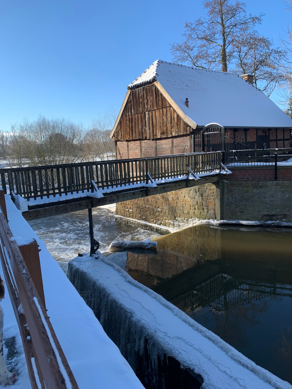 A rustic chalet with wooden elements stands under a clear blue sky, adorned with a layer of fresh snow. A bridge connects the structure to the opposite bank, where water flows gently beneath, partially obscured by snow-covered terrain.