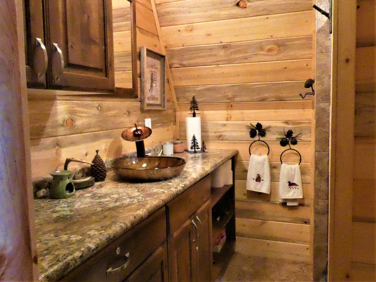 A rustic bathroom is featured, characterized by wooden walls and a granite countertop. A decorative bowl sink sits on the counter, accompanied by pinecones and an art piece. Two towels hang on hooks, while a soap dispenser and a decorative canister are also visible.