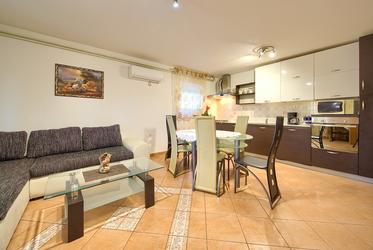 An open-plan living area features a dark sectional sofa adjacent to a glass coffee table. A dining table with light-colored chairs is set for meals. The kitchen, equipped with modern appliances, is in the background, while warm lighting adds a calm ambiance.