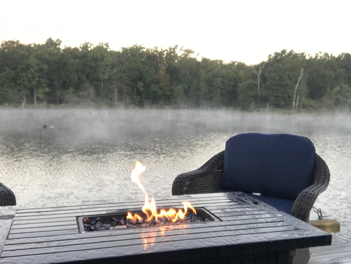 A fire table is situated on a deck, with orange flames dancing above a bed of black rocks. A comfortable blue chair accompanies the table, with a mist rising from the calm water of Vernon Lake, surrounded by a lush, green landscape.