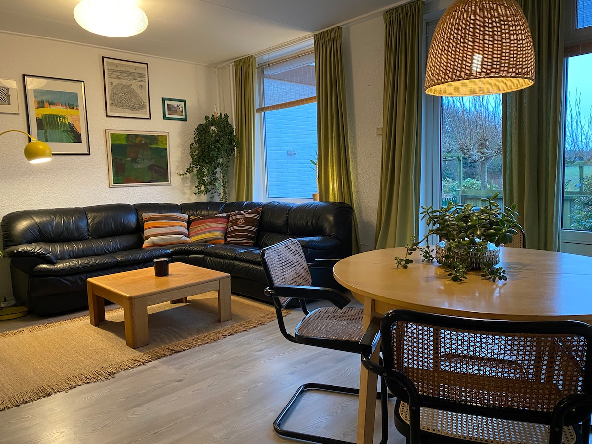 A living area is presented with a dark leather sectional sofa and a round wooden dining table. Green curtains frame the windows, allowing natural light to enhance the space. Wall art adds character, while plants bring a touch of nature indoors.