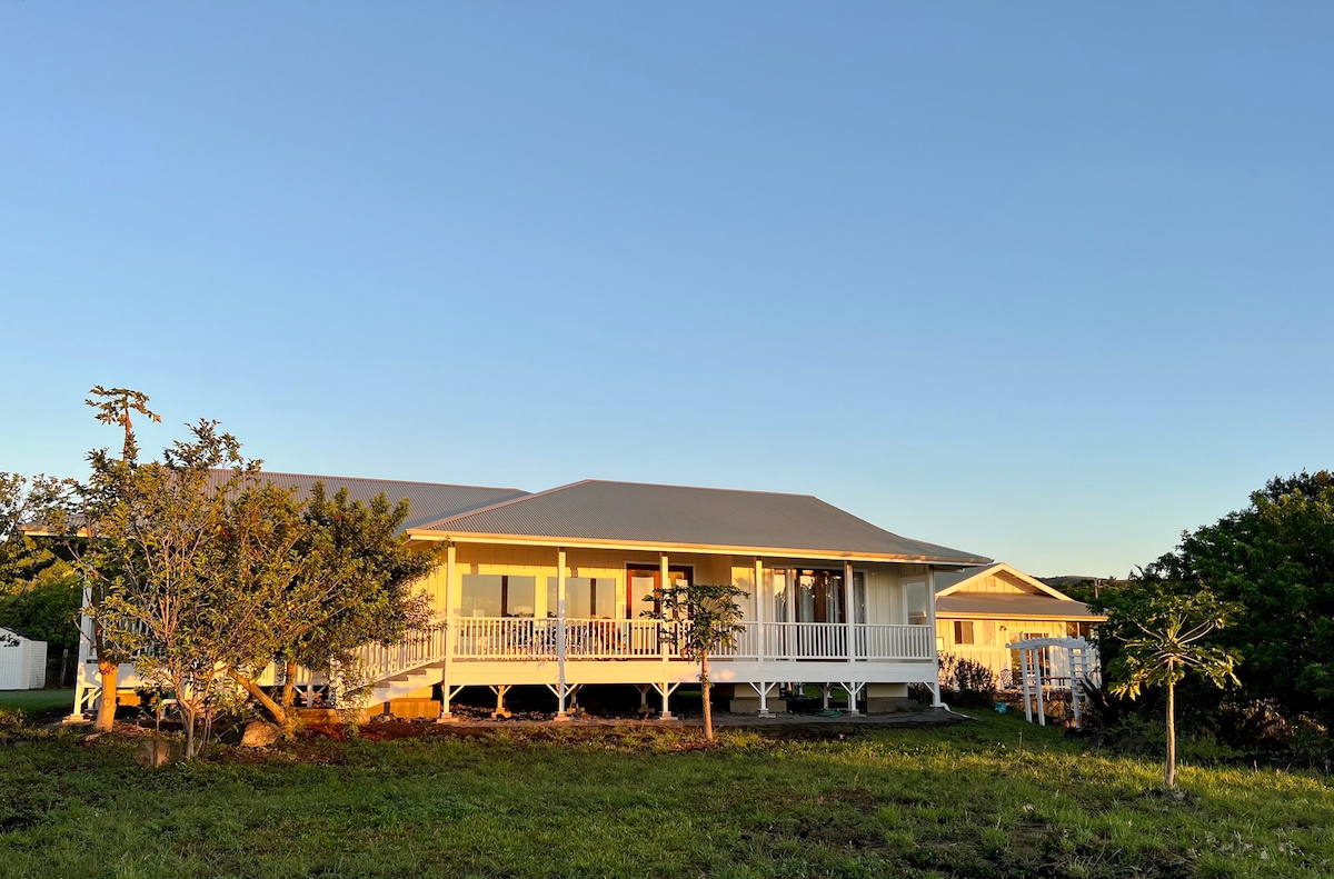 A single-story island home is set against a clear blue sky, featuring a spacious porch that stretches across the front. Lush greenery surrounds the house, enhancing its rural setting. Large windows reflect natural light, offering glimpses into the bright interior.