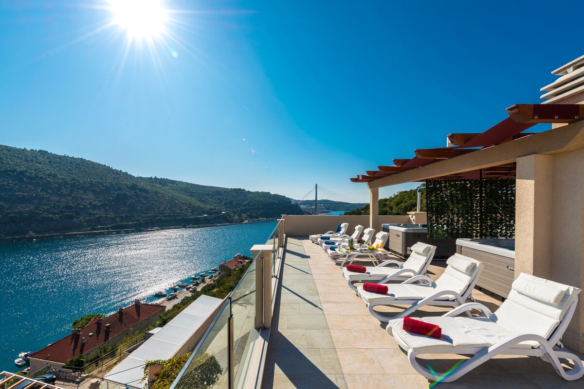 A shared terrace is displayed, featuring sun beds arranged along the edge. A private jacuzzi is positioned nearby, offering expansive views of the sea and surrounding hills. Bright sunlight reflects off the water, creating a serene atmosphere.