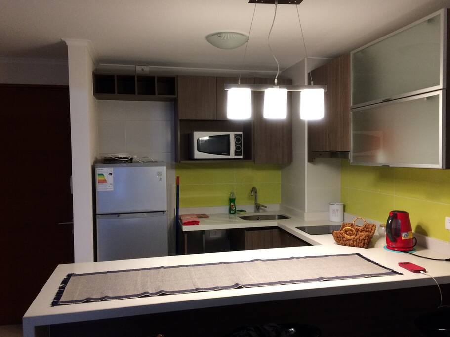 Cocina equipada con Refrigerador, encimera eléctrica Touch, microondas, sillas de bar y hervidor.