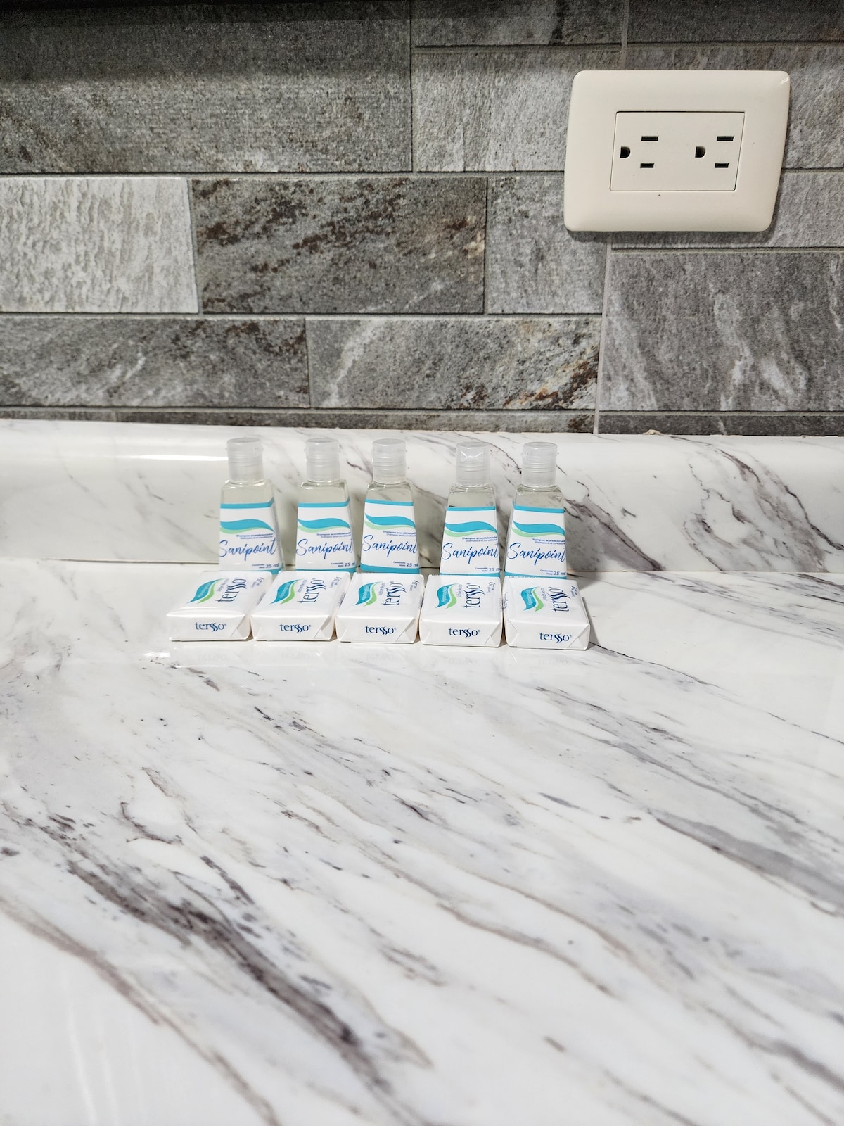 A clean, marble countertop is displayed, featuring a row of five travel-sized toiletries arranged neatly. Each item is labeled with a blue and white design, complementing the natural grey tones of the stone surface. An electrical outlet is visible in the background.