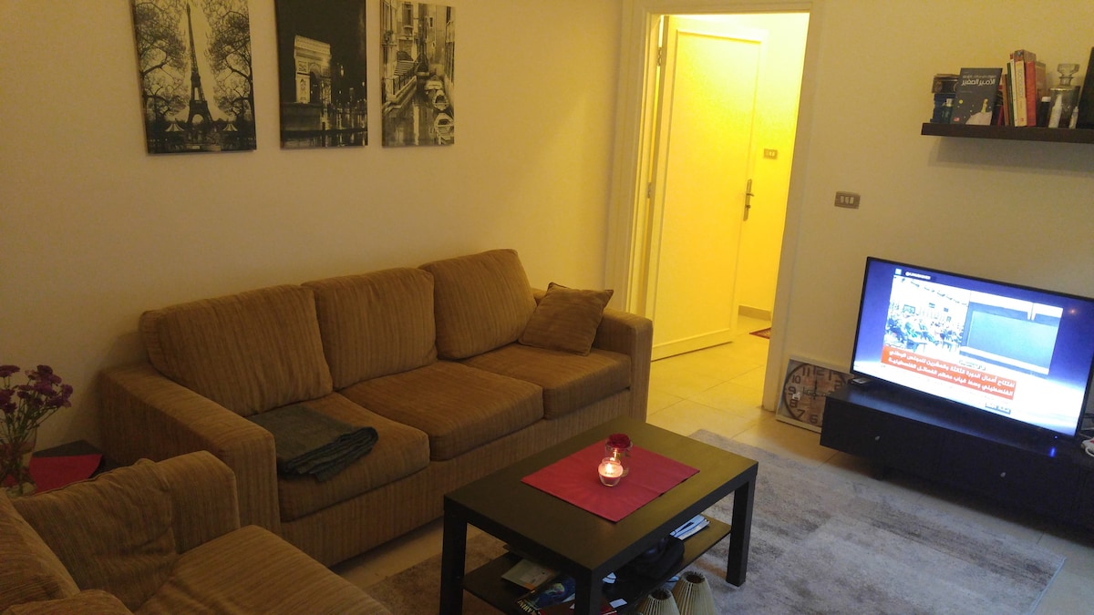 A cozy living area is furnished with a large, comfortable couch and a dark coffee table. A flat-screen TV is positioned on a shelf nearby. The wall displays three black-and-white photographs. A door leading to another room is visible in the background.