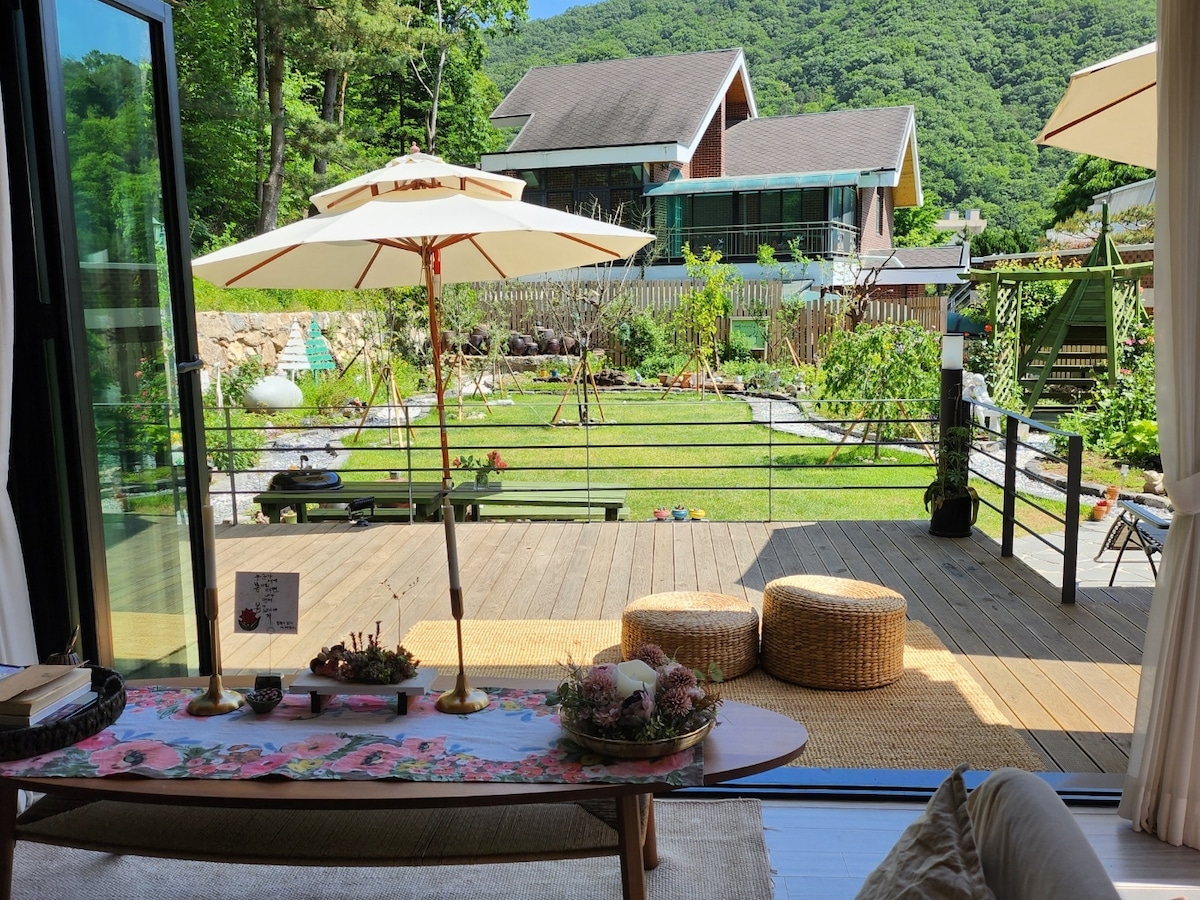 A spacious outdoor area is visible, featuring a wooden deck with two round stools and an umbrella. Lush greenery and decorative plants surround the space, with a backdrop of hills and houses. Natural light fills the area, enhancing the serene ambiance.