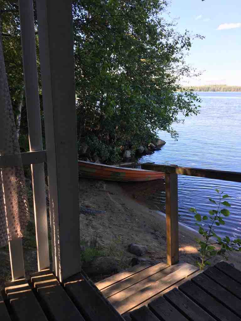 A serene lakeside view is captured from the cabin's terrace, featuring gentle waves lapping against a sandy shoreline. A private rowing boat rests on the beach, surrounded by lush greenery and calm waters reflecting the clear sky.