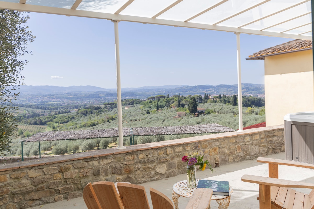 A spacious terrace is presented, featuring wooden chairs and a small table. The view beyond showcases rolling hills with lush greenery, typical of the Tuscan landscape, complemented by a clear blue sky. A hot tub is placed in the corner, enhancing relaxation opportunities.