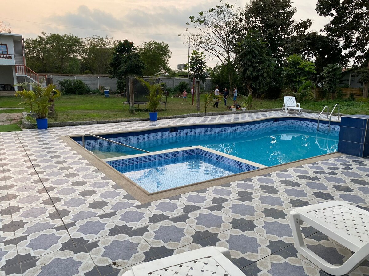 A well-maintained pool area features a spacious swimming pool and a small jacuzzi. Surrounding greenery enhances the peaceful setting. Two lounge chairs are positioned beside the pool on a patterned tile deck, creating an inviting outdoor space for relaxation and enjoyment.