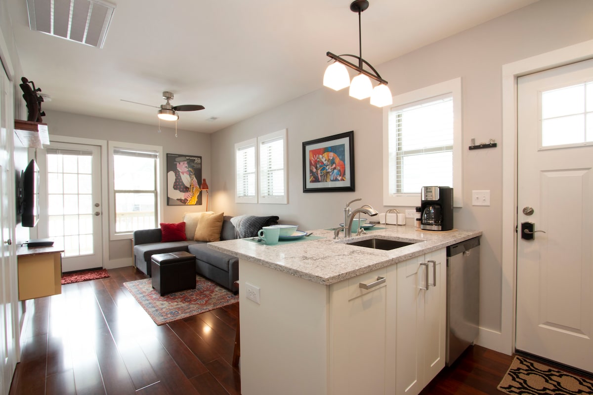 The modern living area features a spacious sectional sofa, a coffee table, and a media stand with a television. A well-equipped kitchen, complete with a granite countertop and coffee maker, is visible. Natural light floods in through two large doors leading to a small outdoor space.