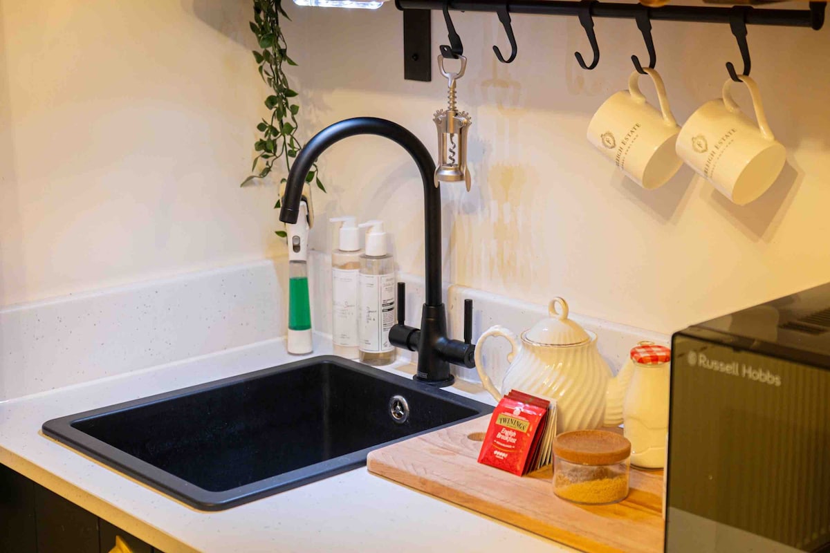 A sleek kitchenette features a black sink with a modern faucet. Above, white mugs hang from a rack, alongside a small dish and a cutting board. Cleaning supplies are neatly arranged on the countertop, while a compact microwave sits nearby.