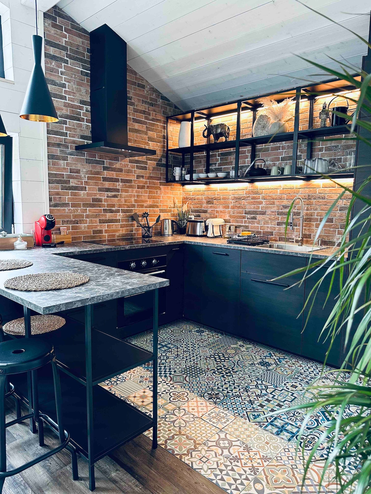 The kitchen features a dark cabinetry and a brick accent wall with warm lighting that illuminates open shelves. A patterned tile floor adds visual interest. Barstools are positioned at a countertop, complementing the modern appliances and stylish layout.