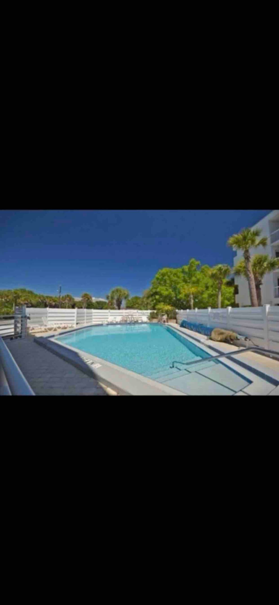 A well-maintained heated pool is surrounded by a clean white deck, with gentle landscaping providing a touch of greenery. The pool steps lead into the inviting water, reflecting the blue sky above and creating a serene atmosphere for relaxation.