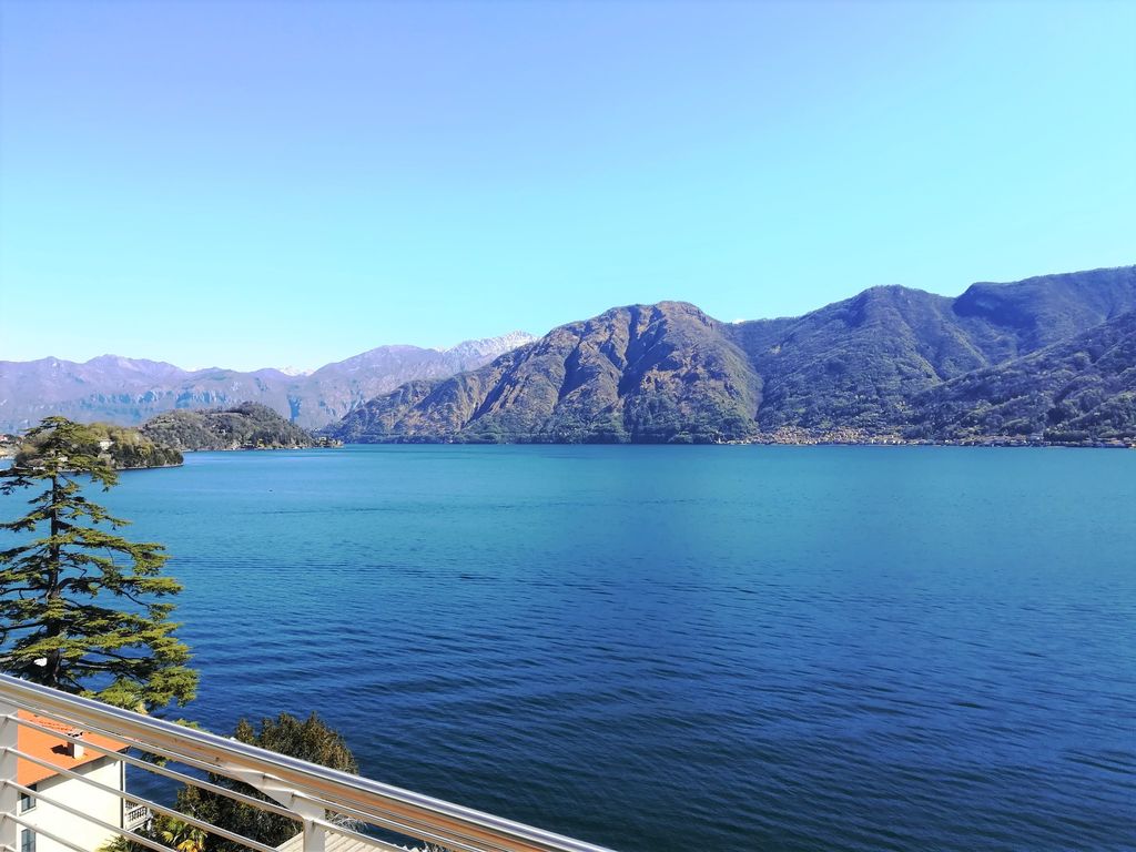 A wide view of the serene Lake Como is showcased, framed by majestic mountains in the background. The clear blue water gently ripples under the sunlight, reflecting the surrounding landscape and creating a peaceful ambiance. Lush trees can be seen along the shoreline.