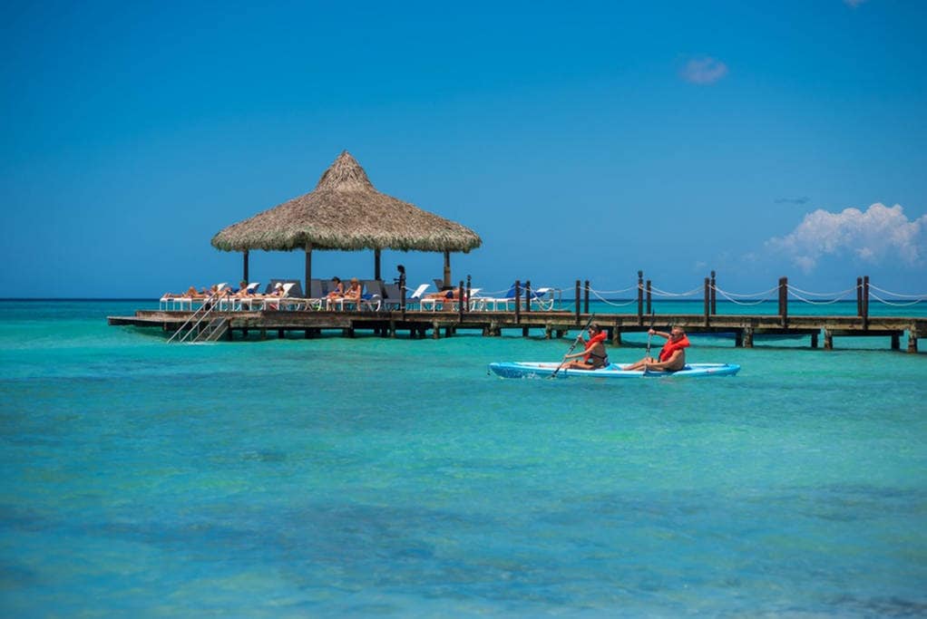 A paddleboard is seen gliding across clear blue waters, with a thatched-roof gazebo on a wooden pier in the background. Sun loungers are positioned around the gazebo, inviting relaxation under a bright sky.