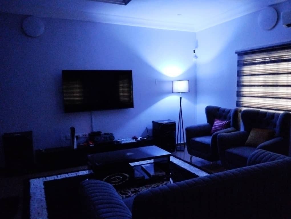 A modern living room features a large wall-mounted television opposite a cozy seating arrangement. Soft lighting creates a calming ambiance, with a floor lamp illuminating one corner. The room includes a plush area rug, enhancing comfort and style.