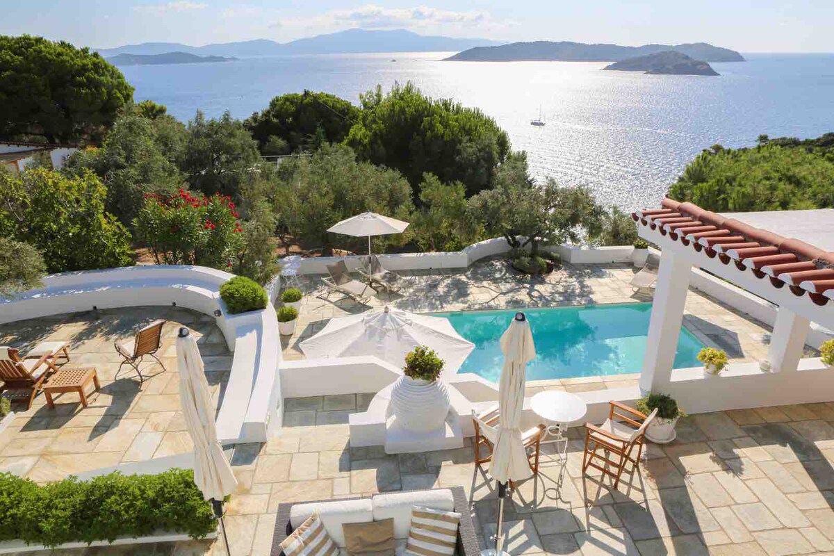 An aerial view showcases a private pool surrounded by a spacious terrace featuring sun loungers and a parasol. Lush greenery lines the property, leading to a sparkling sea with distant islands in the background, creating a serene outdoor setting.