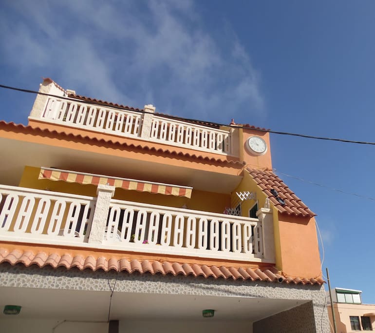 Esta es la fachada de La Casa del Reloj, le decimos así porque si te fijas bien en su fachada colocamos cada año un reloj nuevo que pertenece a la asociación de vecinos. Lo cambiamos porque el agua del mar corroe de manera rápida y agresiva cualquier tipo de superficie metálica.