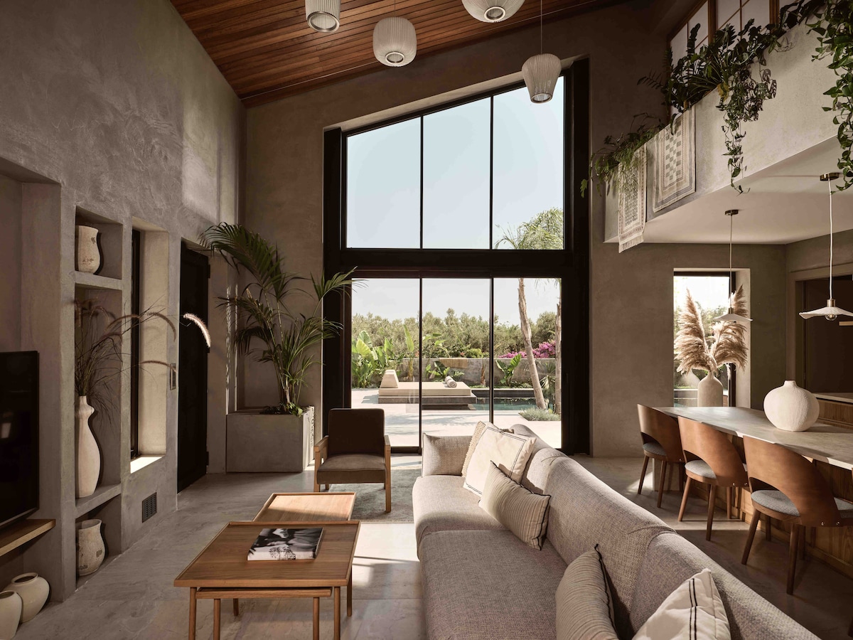 A spacious living area is showcased with high ceilings and large windows that invite natural light. A minimalist sofa is positioned near a wooden coffee table, while plants add a touch of greenery. The outdoors is visible, featuring a pool and lush garden.