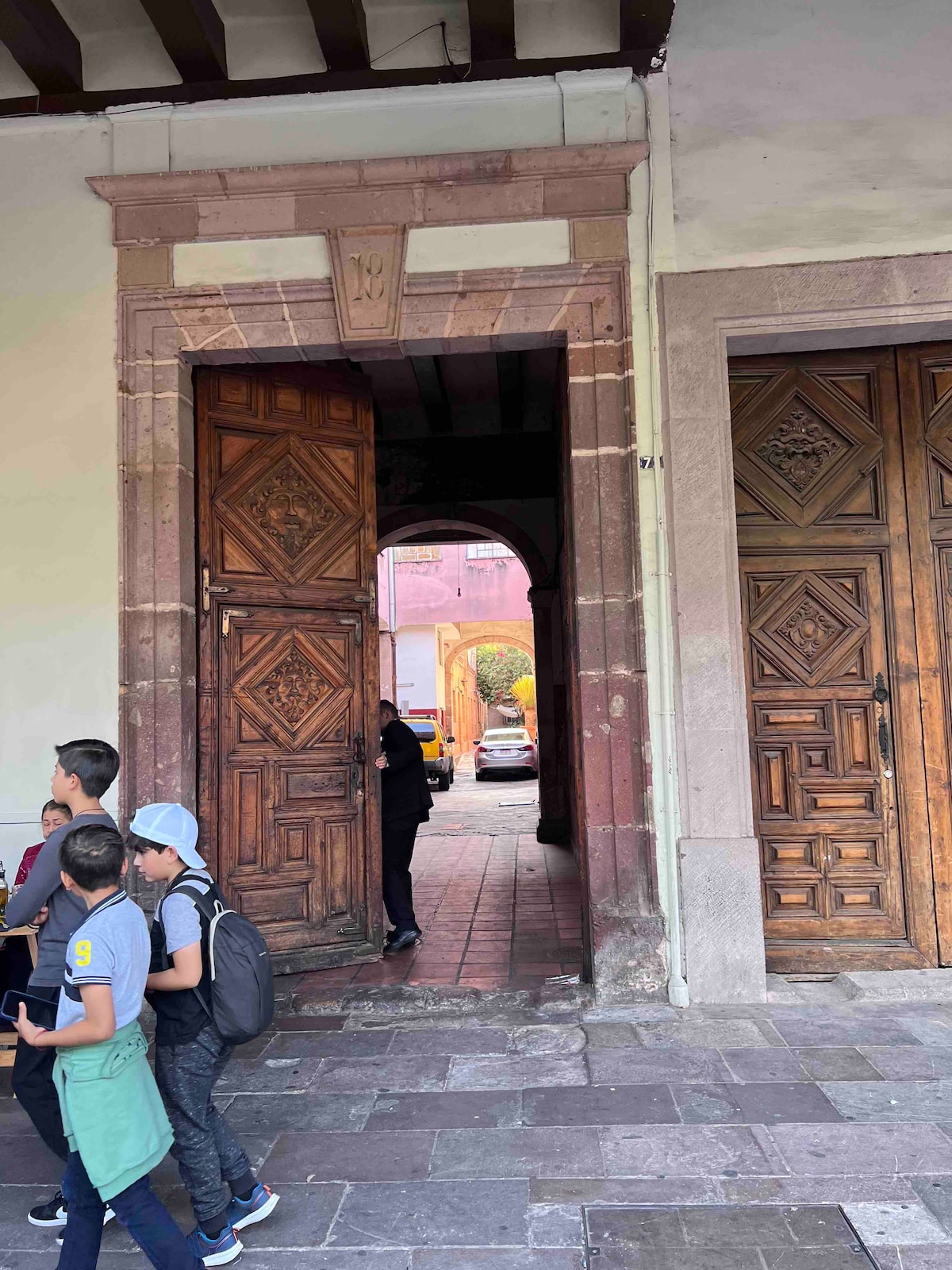A pair of intricately carved wooden doors is shown, leading to a spacious entrance. The path is bordered by stone flooring, and individuals are observed walking past the doorway. Soft natural light filters through, highlighting the craftsmanship of the door designs.