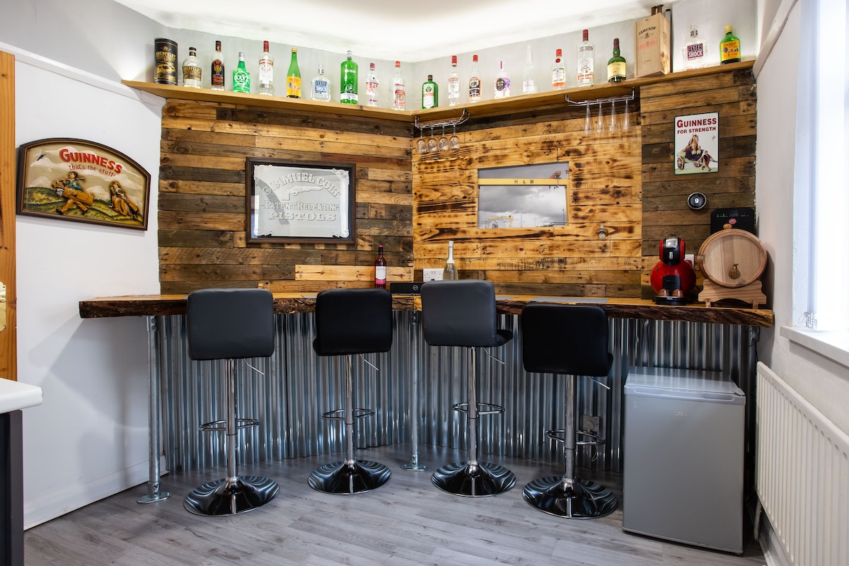 A cozy bar area features a wooden counter supported by metallic stands and equipped with high black chairs. The wall is adorned with a variety of vintage bottles and framed memorabilia, providing a rustic charm. A small refrigerator is positioned nearby for convenience.