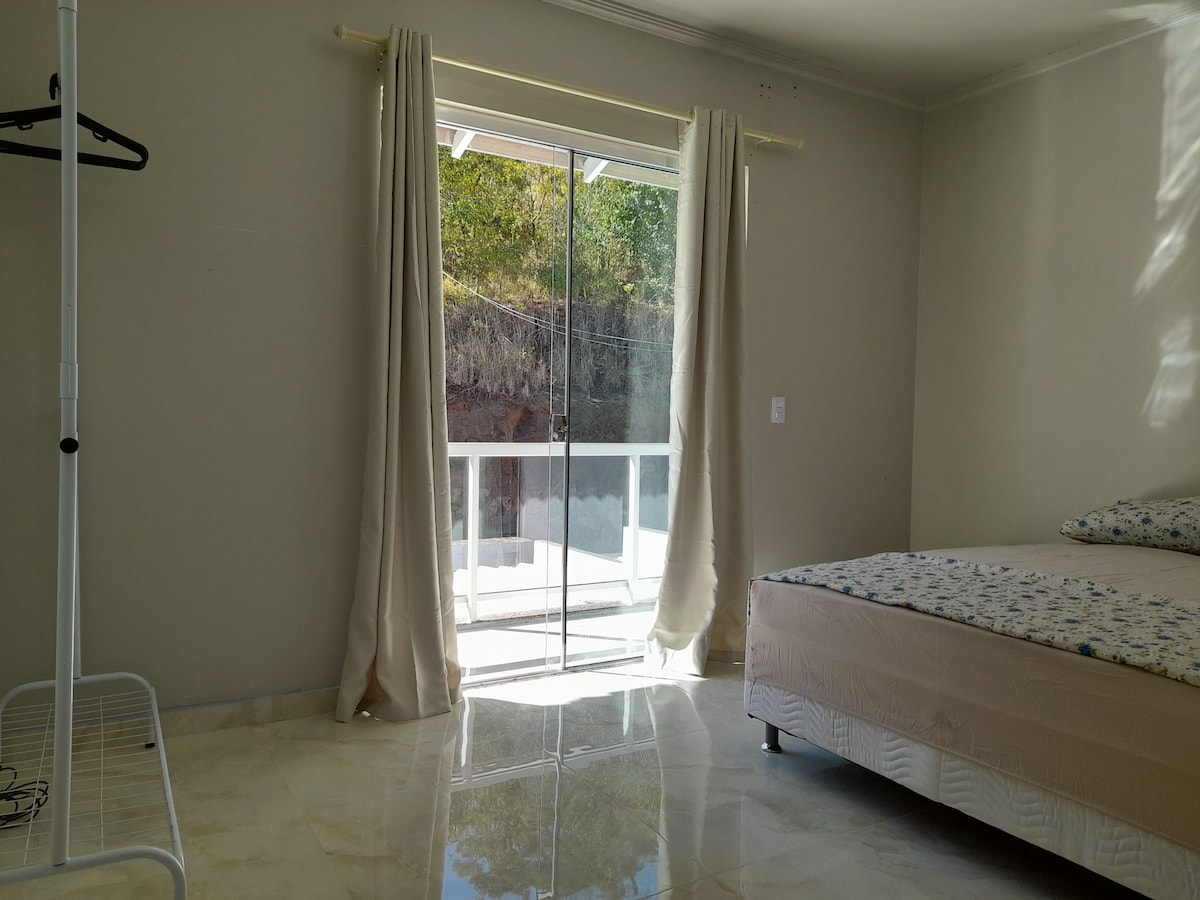 A bright sleeping area features a spacious bed with a patterned mattress. Light curtains frame a sliding glass door that opens to a balcony, allowing natural light to illuminate the marble floor. A clothing rack is positioned beside the door, adding functionality to the space.