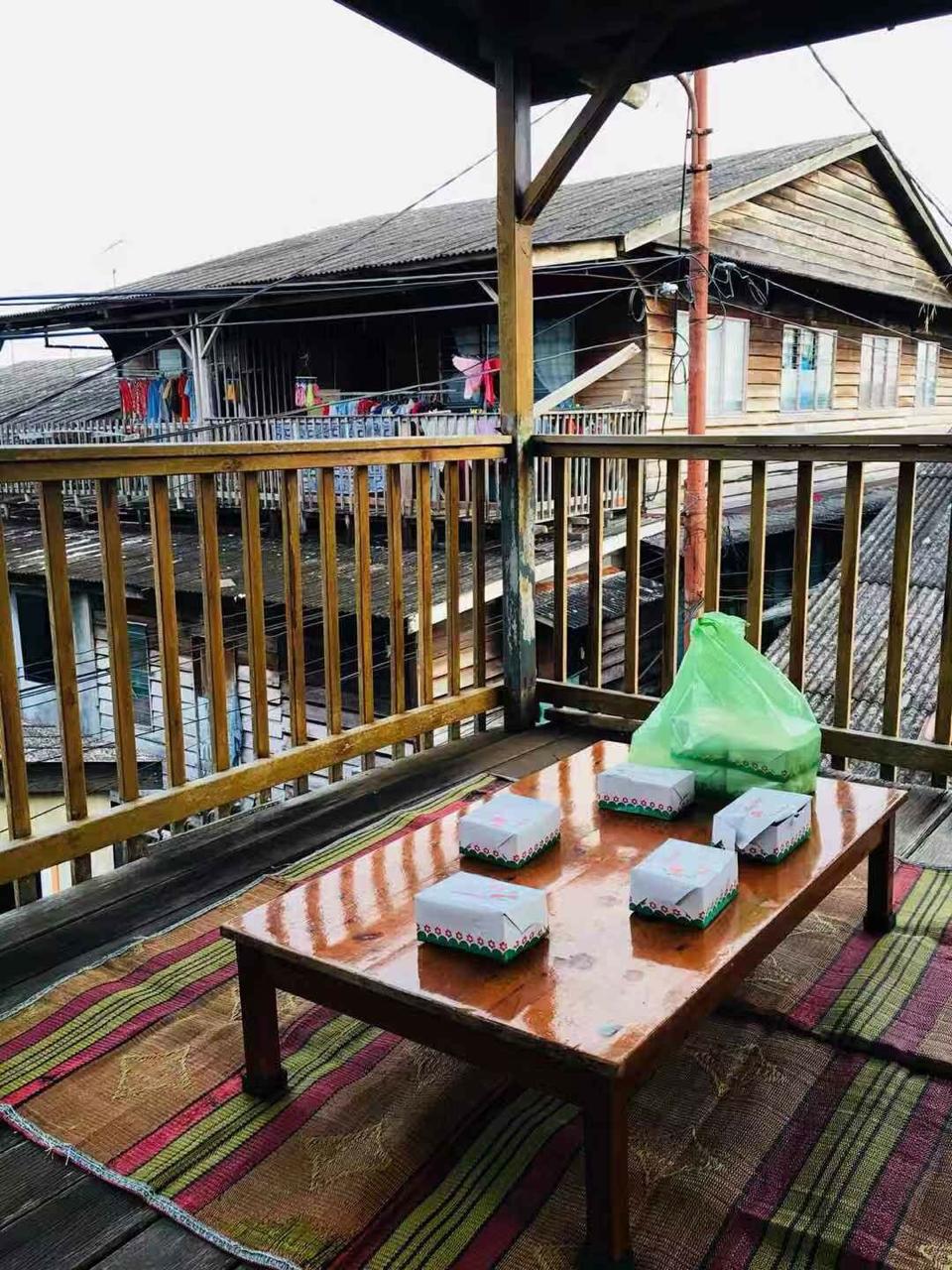 A wooden balcony features a table set with six takeout boxes, offering a view of neighboring wooden structures. The floor is covered with a woven mat, and a green plastic bag rests on the table. Clotheslines with colorful garments hang in the background.