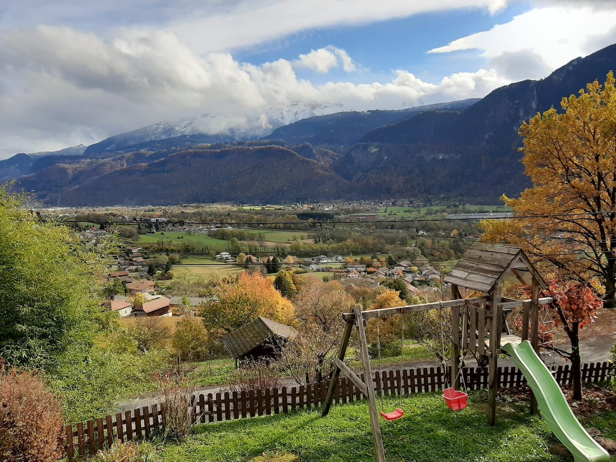 A panoramic view of mountains is captured from the terrace, showcasing a colorful landscape of trees and fields. A playground with a slide and swings is visible in the foreground, along with a wooden fence that encloses the area.
