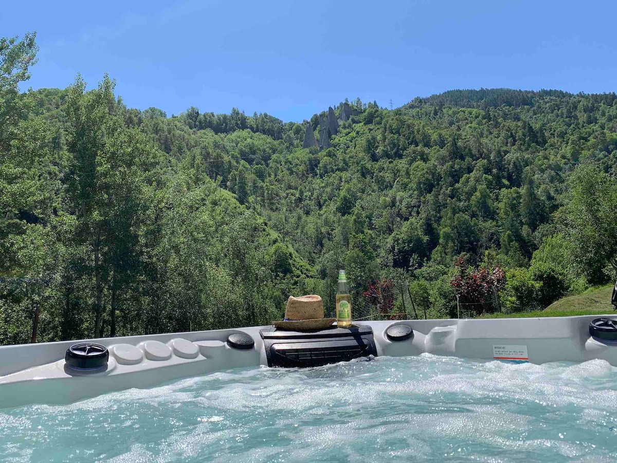 The image depicts a relaxing hot tub surrounded by lush green trees and scenic hills. A straw hat and a bottle of drink are placed on the edge, inviting guests to unwind while enjoying the natural landscape. Clear blue skies provide a bright backdrop.