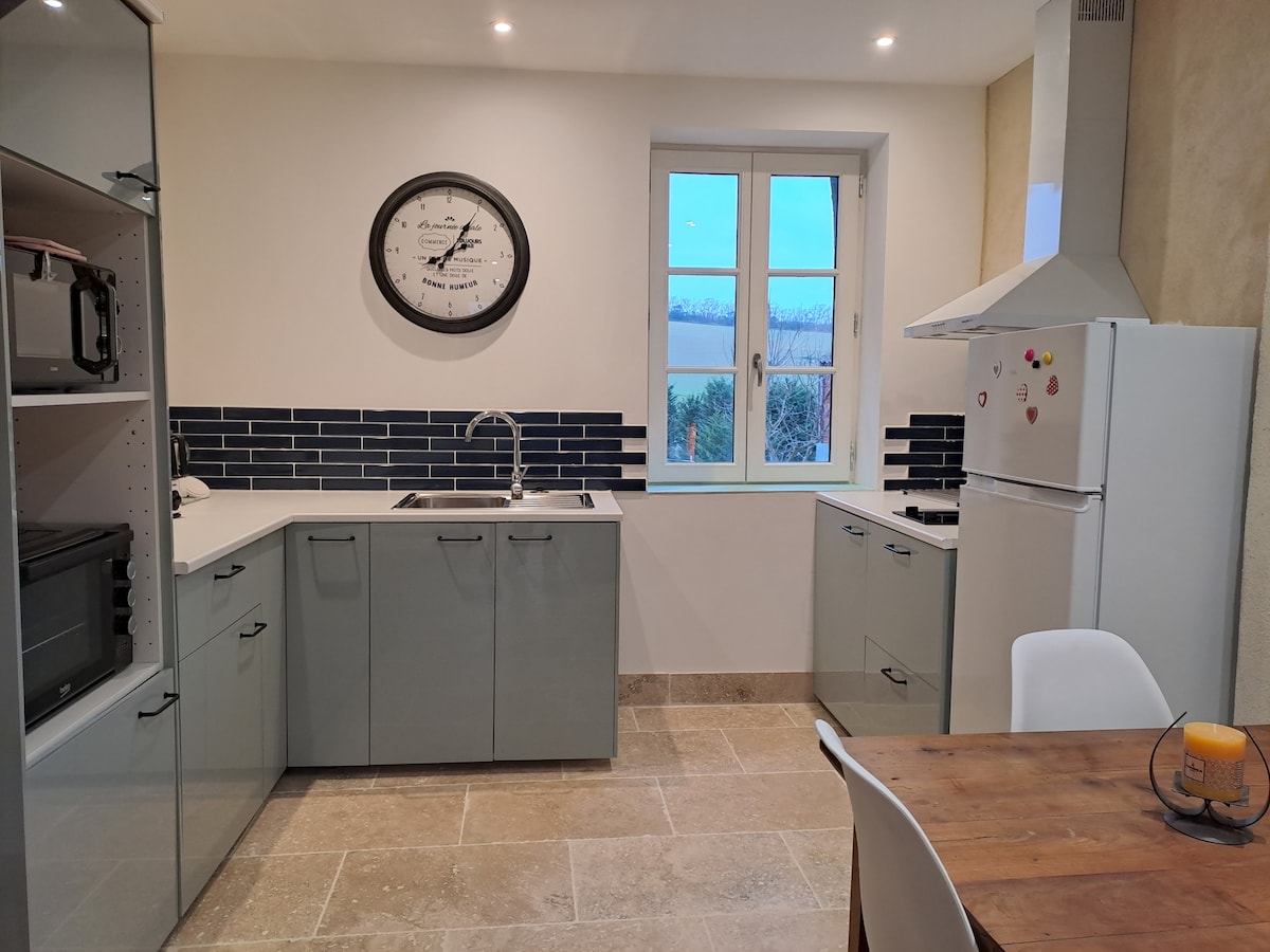 A modern kitchen is presented with sleek cabinets and a neutral color palette. A functional layout includes an oven, microwave, and refrigerator. Natural light enters from a double window, while a round wooden table and chairs provide an inviting space for meals.