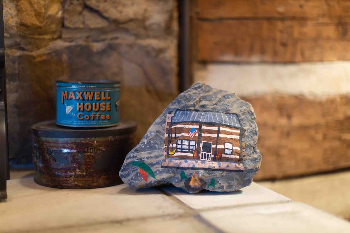 A rustic decorative arrangement is displayed, featuring a painted rock depicting a log cabin. Nearby, a vintage tin of Maxwell House coffee adds character to the setting, complemented by wooden accents in the background.