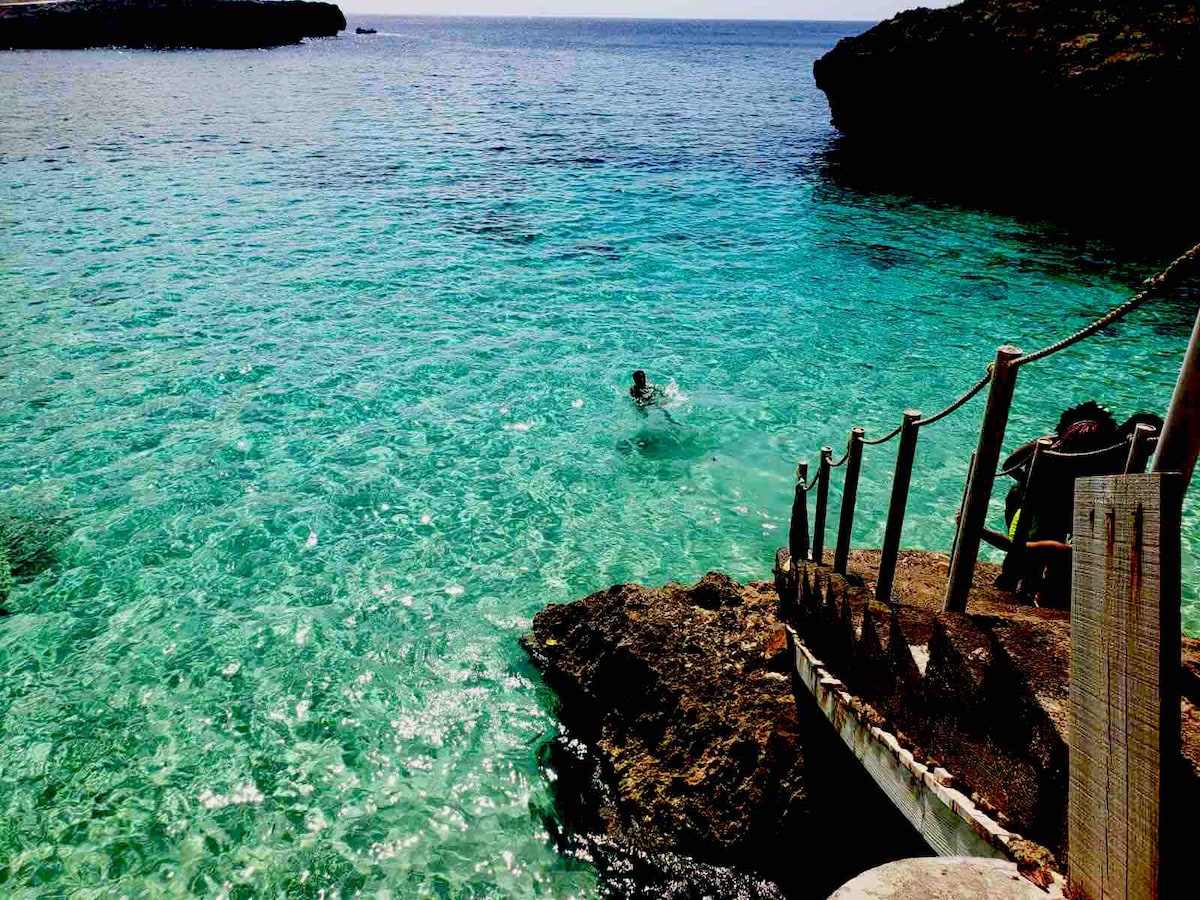 A vibrant turquoise sea glistens under sunlight, revealing varying depths of clear water. Rocky formations frame the scene, with a rustic staircase leading down to the water's edge. A swimmer can be seen among the gentle waves, enjoying the calm aquatic environment.