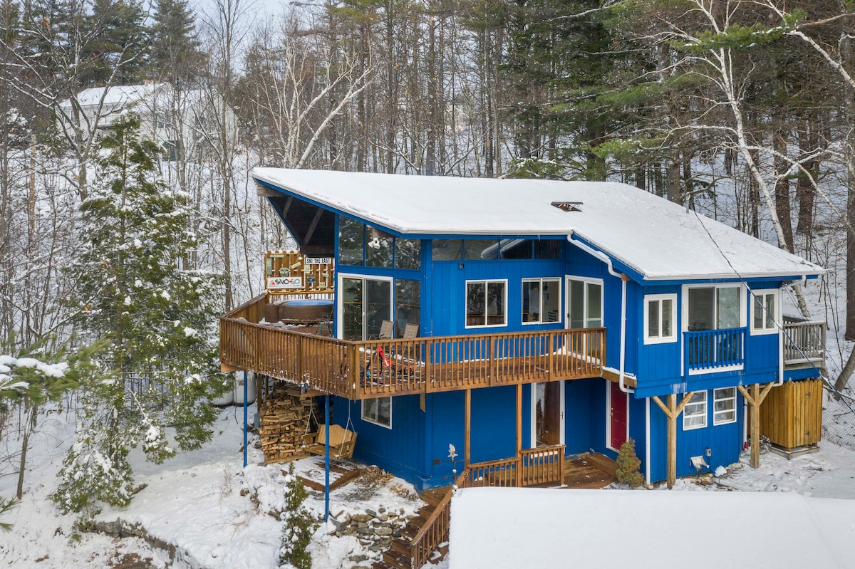 The chalet's vibrant blue exterior is complemented by snow-covered details. A spacious deck wraps around the home, featuring outdoor seating. Surrounding pine trees create a natural backdrop, while the sloped terrain indicates the mountain setting. Stacks of firewood are neatly arranged beside the entrance.
