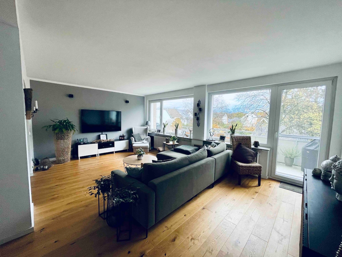 A spacious living area features large windows that allow natural light to brighten the room. Comfortable seating arrangements include a sectional sofa and a chair. A minimalist entertainment unit holds a flat-screen TV, while potted plants add a touch of greenery to the ambiance.