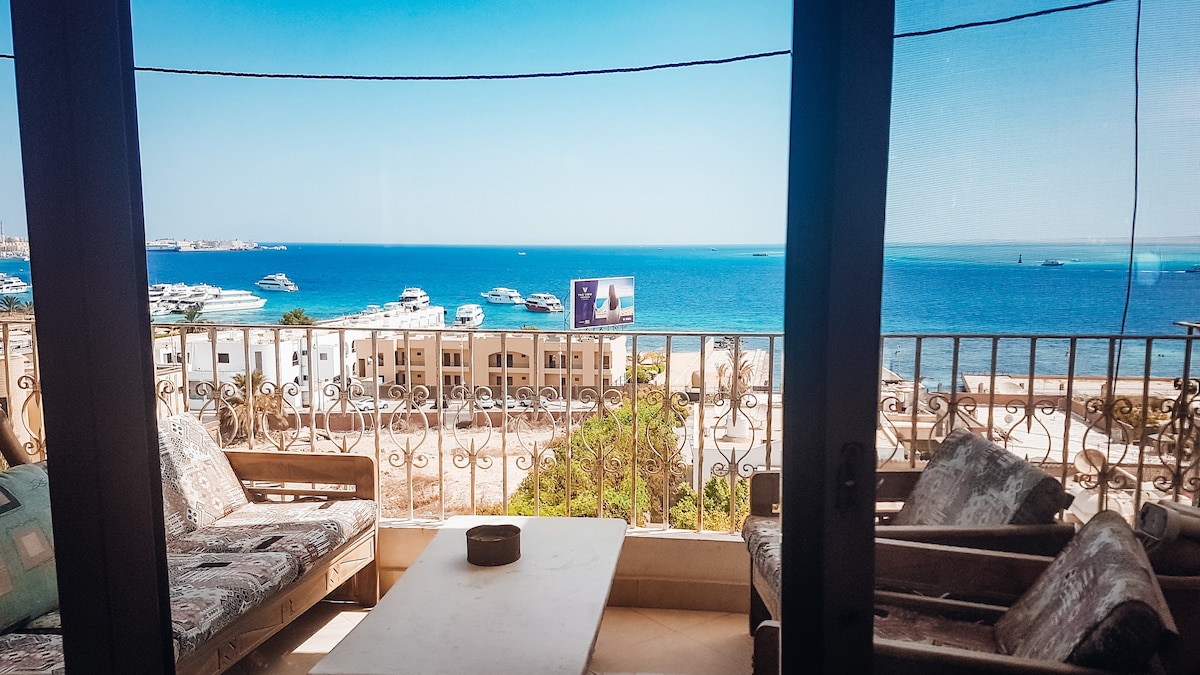 The balcony offers a panoramic view of the sea, with boats visible on the water. Comfortable seating arrangements, including a rustic table, provide an inviting space to relax while enjoying the scenery. Natural light enhances the open atmosphere.