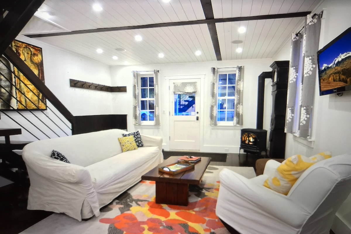 A welcoming living area features light-colored sofas arranged around a coffee table. Bright artwork adorns the walls, complemented by natural light from two windows. A cozy wood stove and a mounted television are present, with modern lighting illuminating the space.