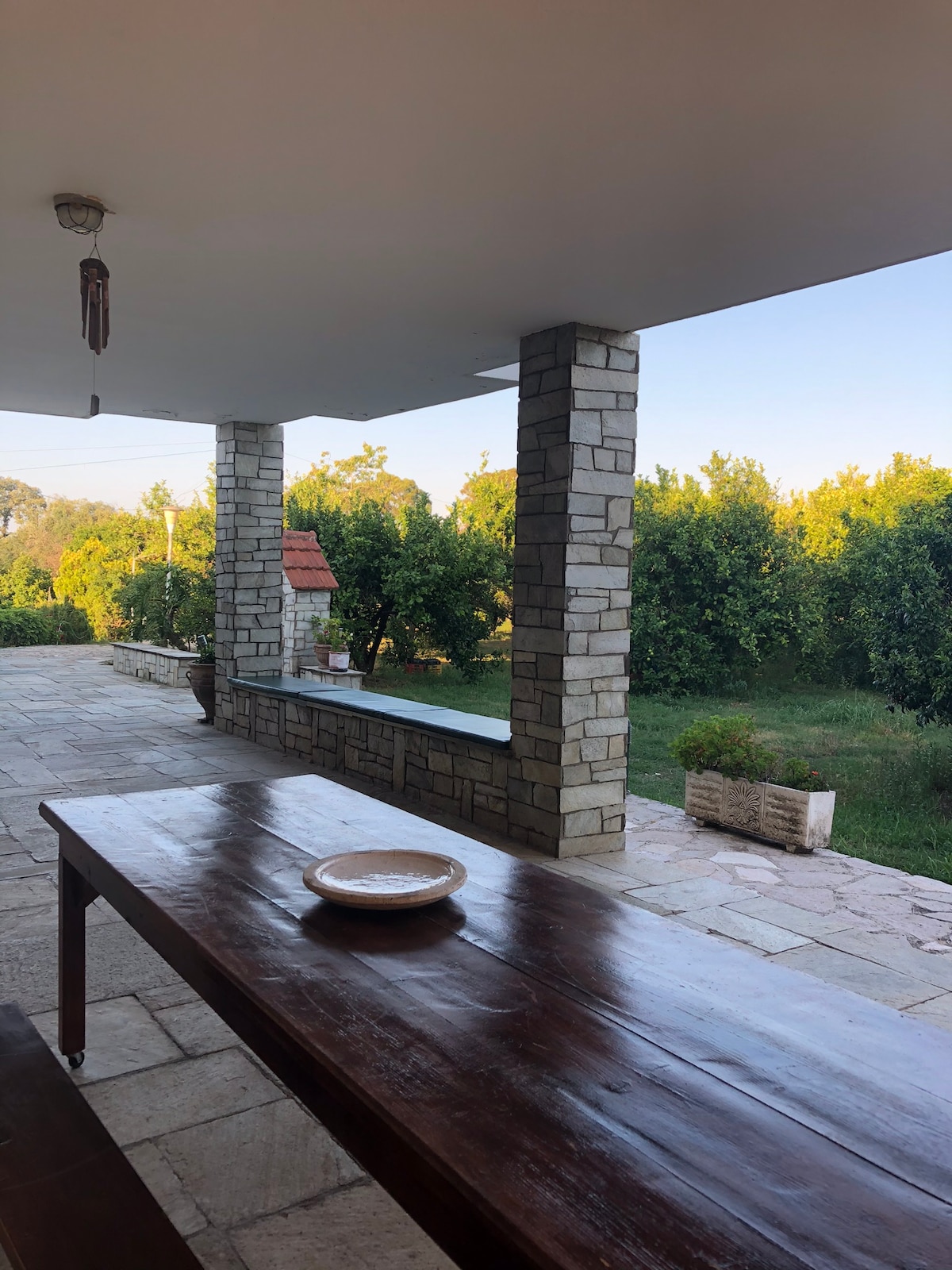 A spacious stone terrace features a large wooden table set against a backdrop of vibrant greenery. Stone columns frame the space, providing a blend of indoor comfort and outdoor tranquility. Natural light filters in, enhancing the fresh, open atmosphere of the area.