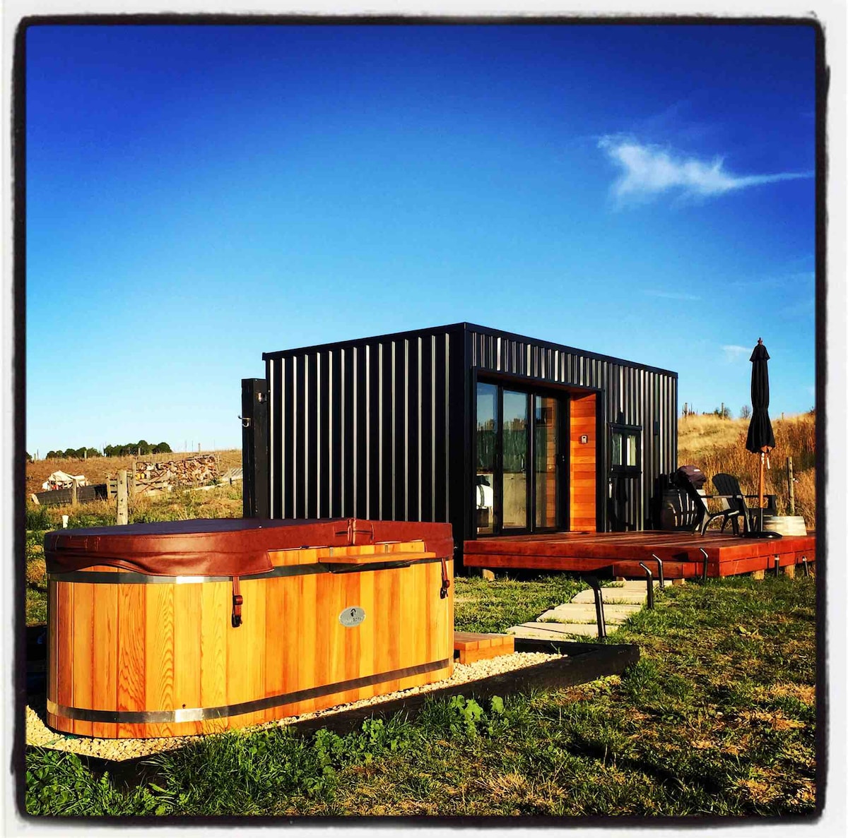 A wooden hot tub is positioned next to a modern tiny house with sleek black siding. A wooden deck extends from the house, featuring two chairs and a table. The clear blue sky and surrounding natural landscape provide a serene backdrop.
