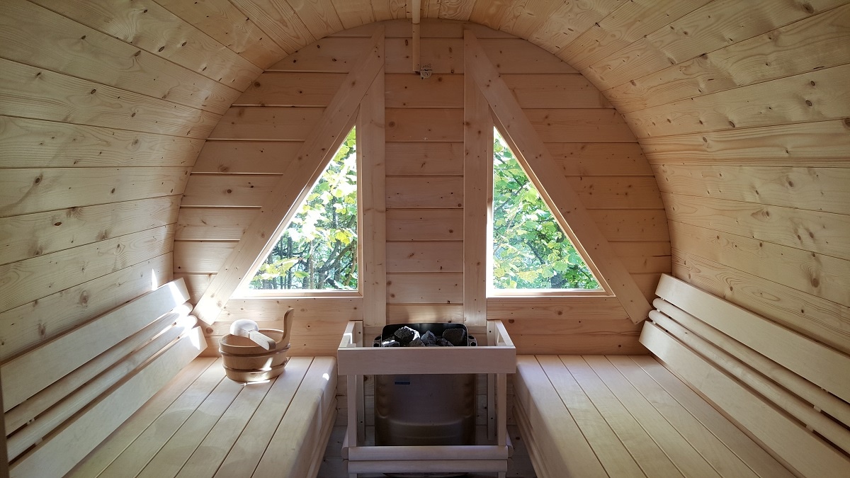 The sauna features a cozy wooden interior with triangular windows allowing natural light and views of greenery. A stone heater is centered, surrounded by wooden benches, creating a serene space for relaxation.