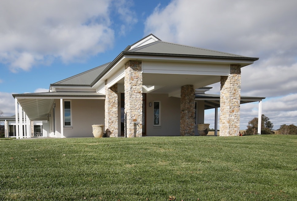 The exterior of the Homestead is showcased, featuring a contemporary design with stone-clad pillars. Surrounding green lawns extend toward the horizon under a partly cloudy sky, emphasizing the property's spacious layout and tranquil rural setting.